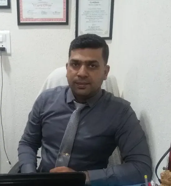 A physiotherapist in a shirt and tie seated at his desk at the clinic, with the lower corners of two framed certificates visible on the wall behind him.
