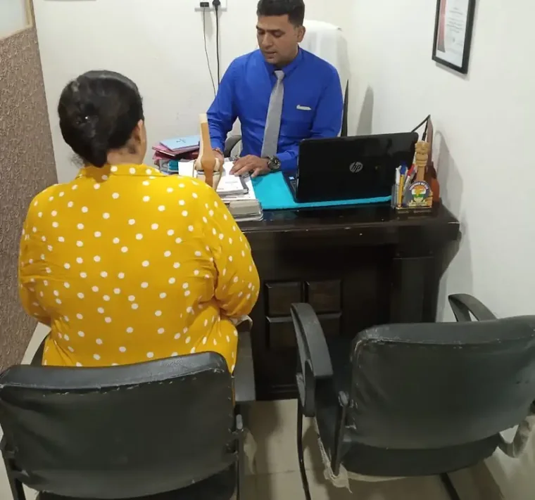 A consultation room: one person in a blue shirt and tie sits behind a dark wooden desk with a laptop and files, another sits opposite with their back to the camera; a framed certificate hangs on the wall behind.