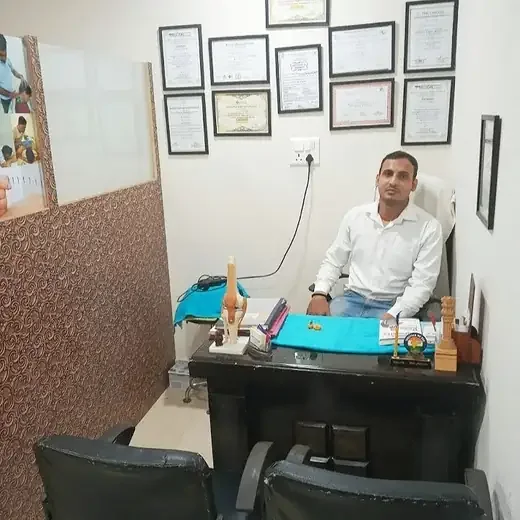 A desk area at the clinic: a dark desk with a teal mat, an anatomical knee model and small awards, a person in a white shirt seated behind it, and a wall of framed certificates above.