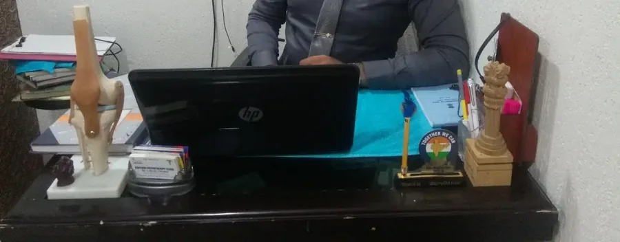 Close view along the clinic desk: an anatomical model of a knee joint, an open laptop, a card holder, pens and small wooden desk ornaments, with someone seated behind the laptop out of frame.